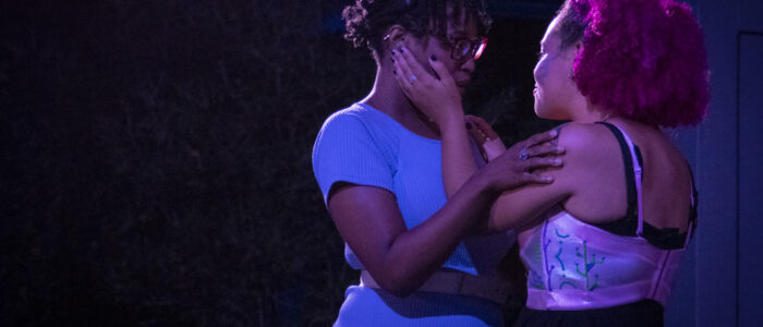 Two actors are holding eachother in an embrace. One, with pink hair, is holding the other's face, while the other, who is wearing blue, is holding the pink haired shoulders.