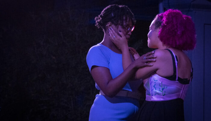 Two actors are holding eachother in an embrace. One, with pink hair, is holding the other's face, while the other, who is wearing blue, is holding the pink haired shoulders.